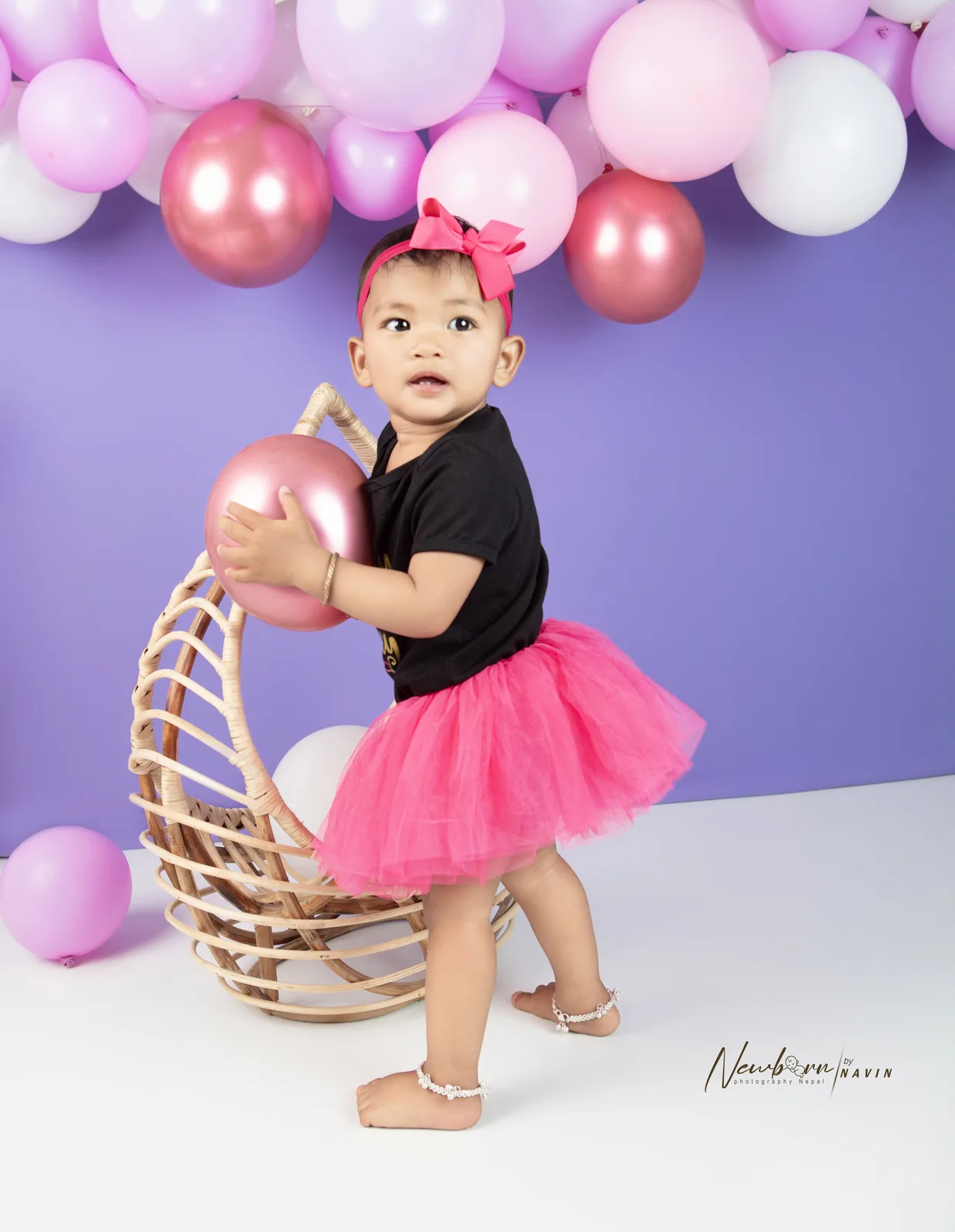 One-year-old girl in a pink tutu and headband holding a pink balloon beside a rattan hanging chair, styled for a birthday photography session against a purple backdrop.
