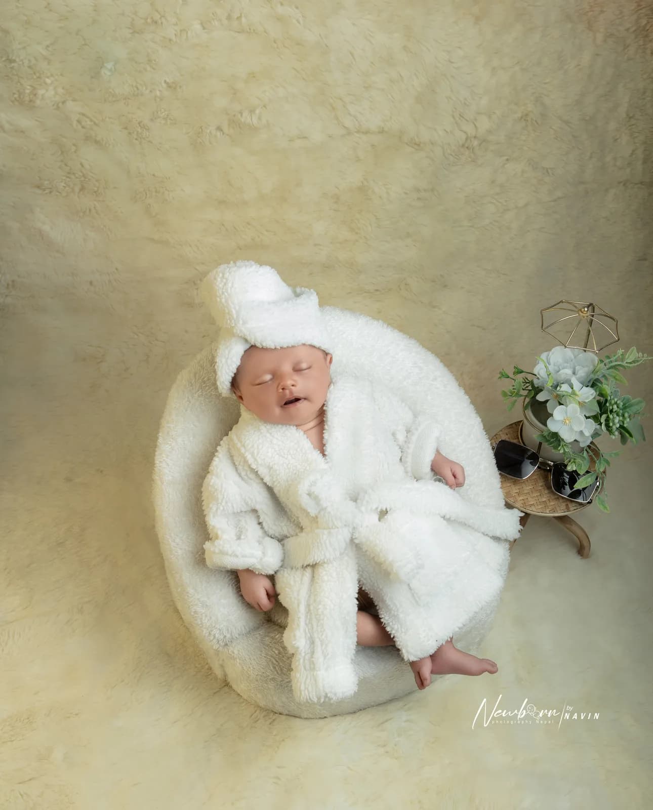 Newborn baby dressed in a miniature white spa robe and towel wrap, posed sitting in a round fur chair beside sunglasses and a small flower arrangement.