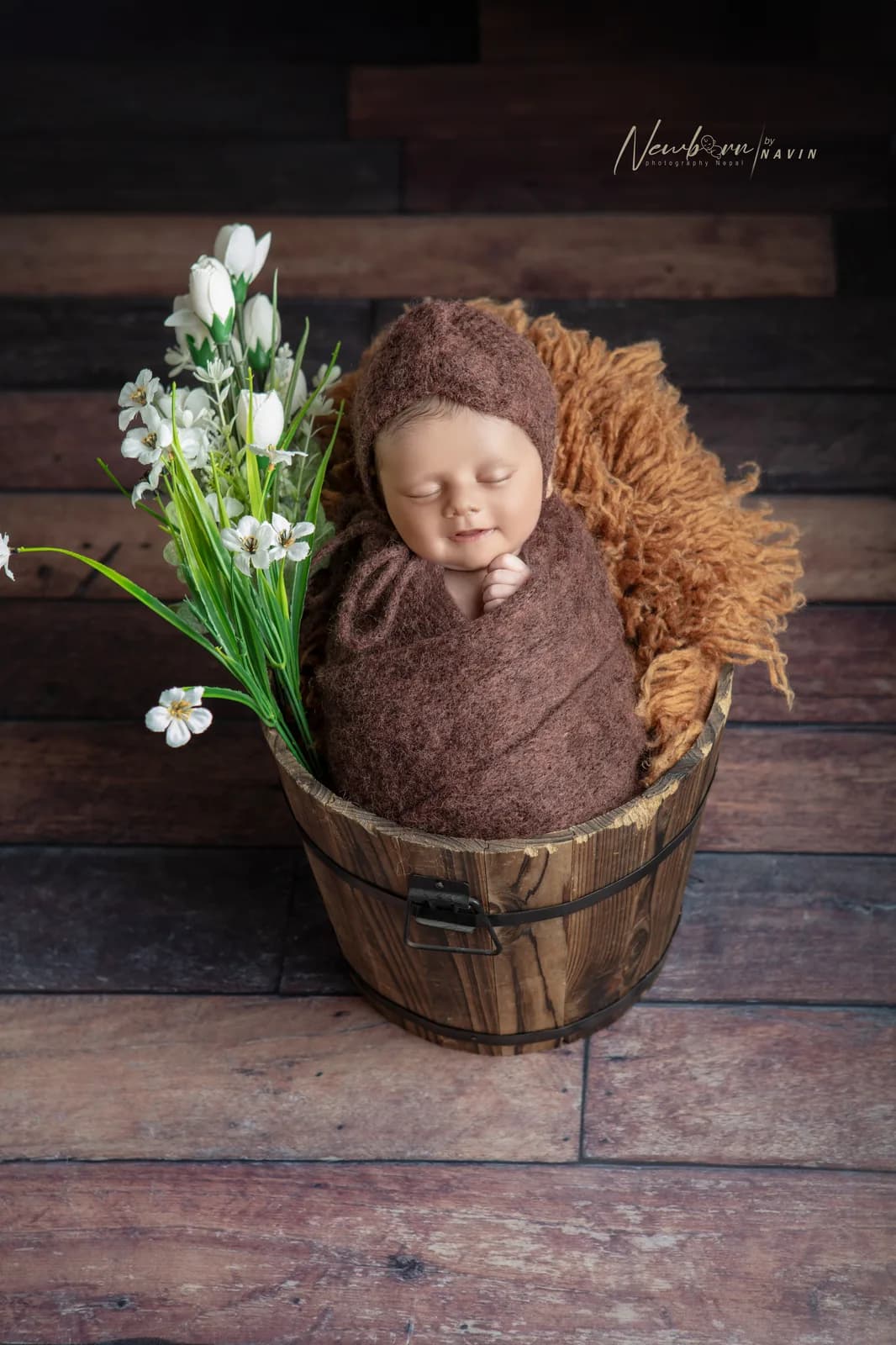 Smiling newborn baby wrapped in a brown knit wrap and matching bonnet, sitting inside a wooden bucket prop with white flowers, against a wood-plank backdrop.
