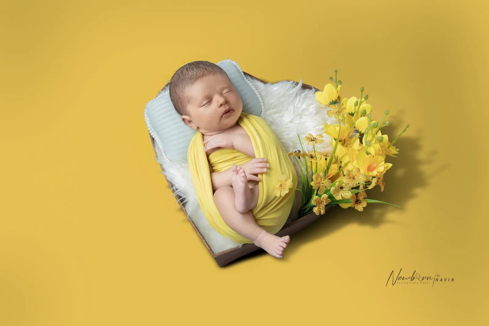 Newborn baby wrapped in a yellow wrap, sleeping on a small wooden bed prop against a warm yellow studio backdrop with yellow flowers.